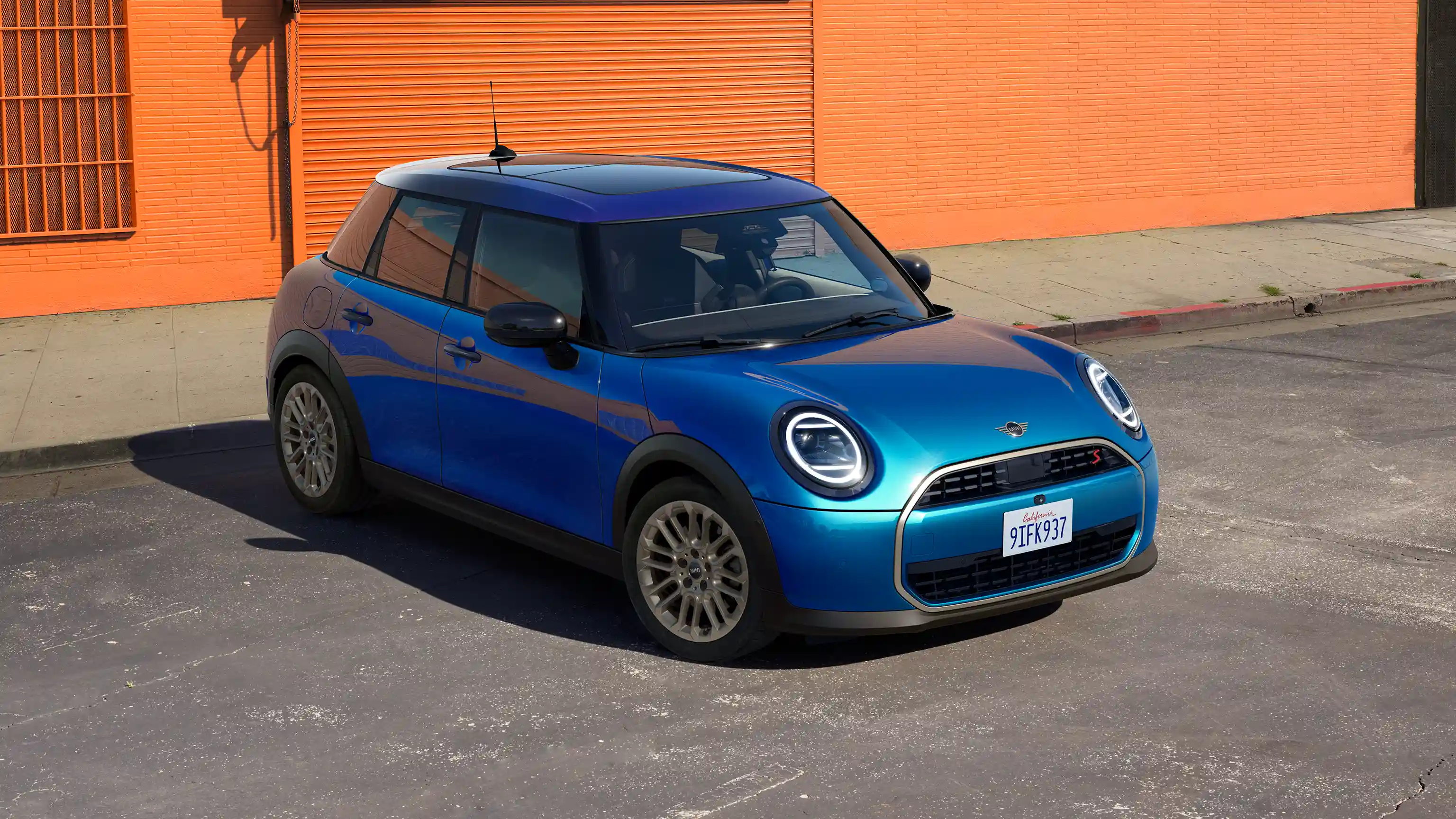 Vista frontal de um MINI Cooper de 5 portas, em Icy Sunshine Blue, com tejadilho  Multitone Blue, estacionado na rua com uma parede cor de laranja em segundo plano