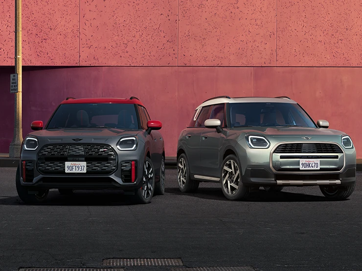 Vista frontal do MINI Countryman JCW em Legend Grey e Red Roof e do MINI Countryman em Smokey Green e tejadilho Vibrant Silver parados ao lado um do outro.