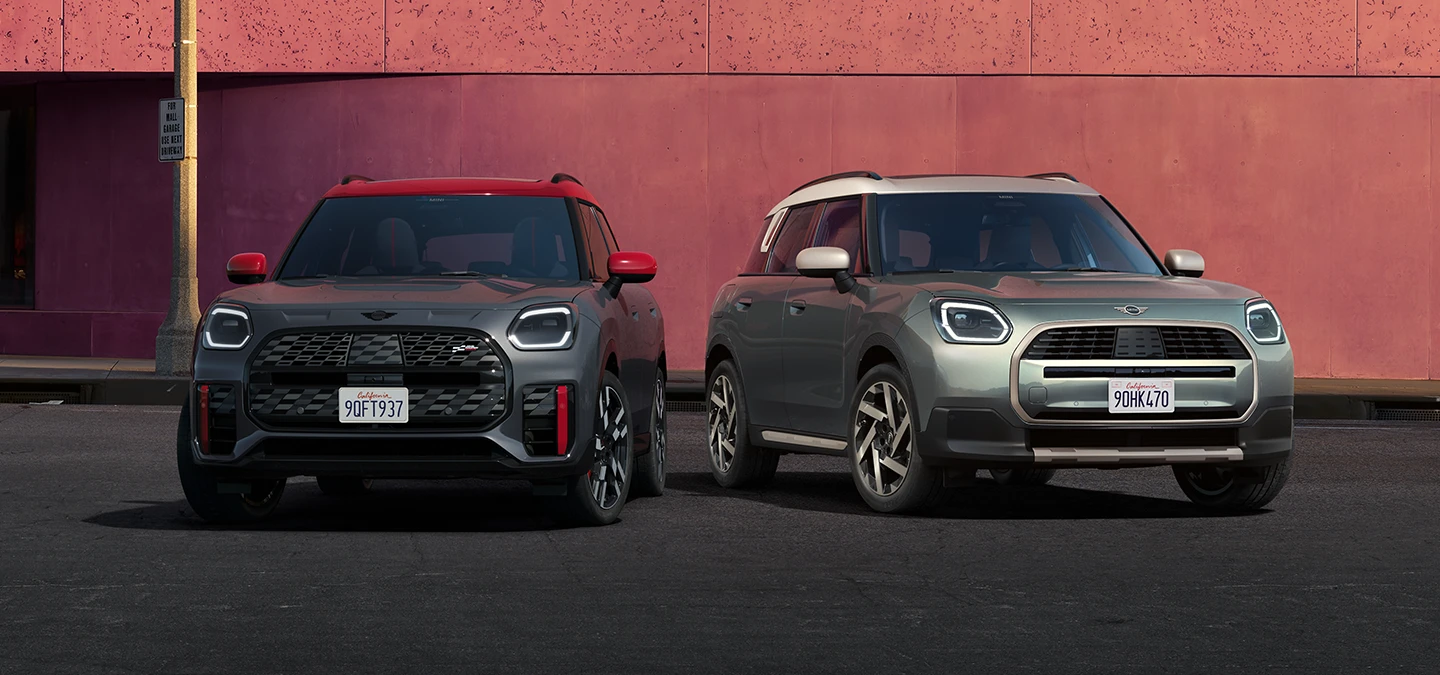 Vista frontal do MINI Countryman JCW em Legend Grey e Red Roof e do MINI Countryman em Smokey Green e tejadilho Vibrant Silver parados ao lado um do outro.