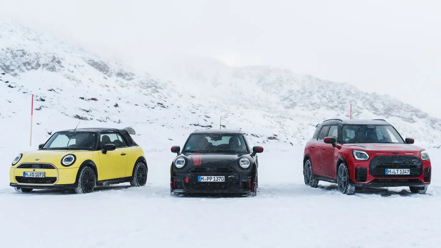 MINI Cooper Descapotável em Sunny Side Yellow, MINI John Cooper Works em Midnight Black II e MINI Countryman Cooper em Chilli Red – estacionados numa estrada de montanha com neve, vista frontal.