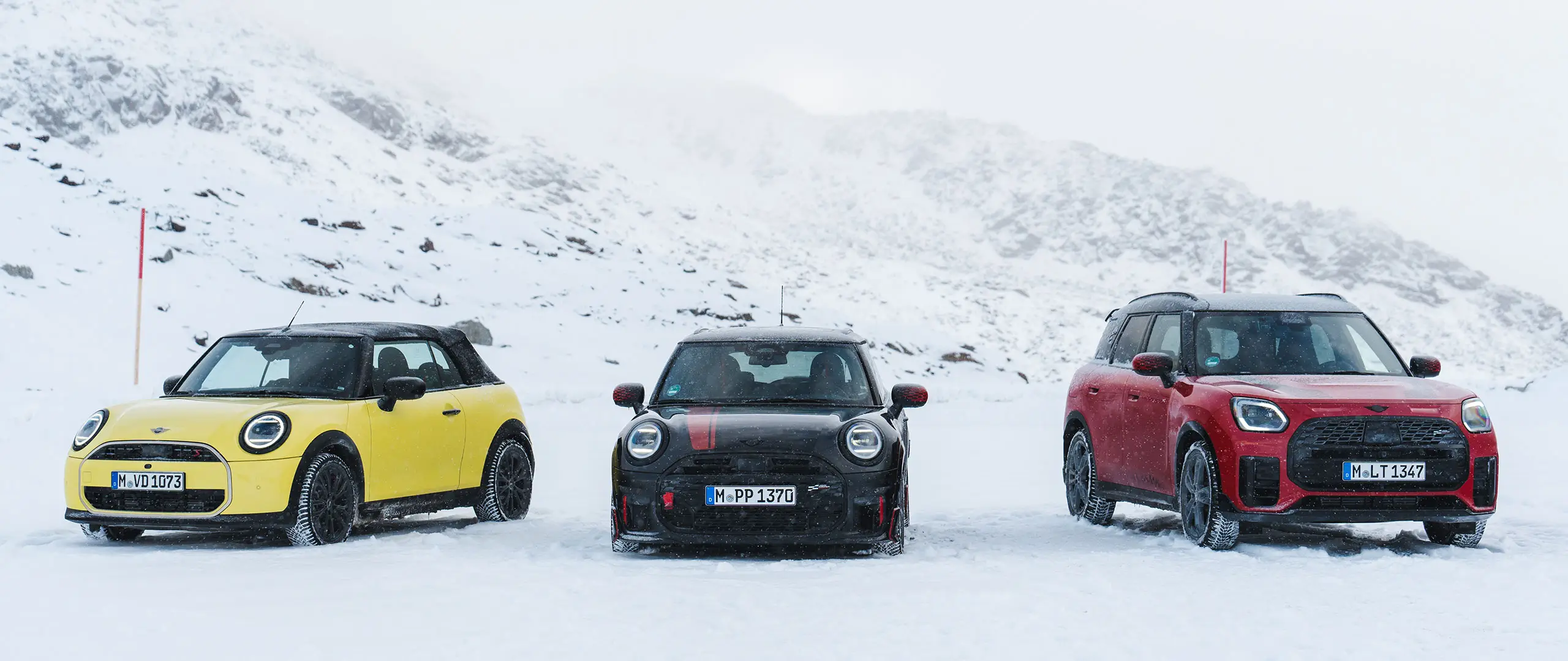 MINI Cooper Descapotável em Sunny Side Yellow, MINI John Cooper Works em Midnight Black II e MINI Countryman Cooper em Chilli Red – estacionados numa estrada de montanha com neve, vista frontal.