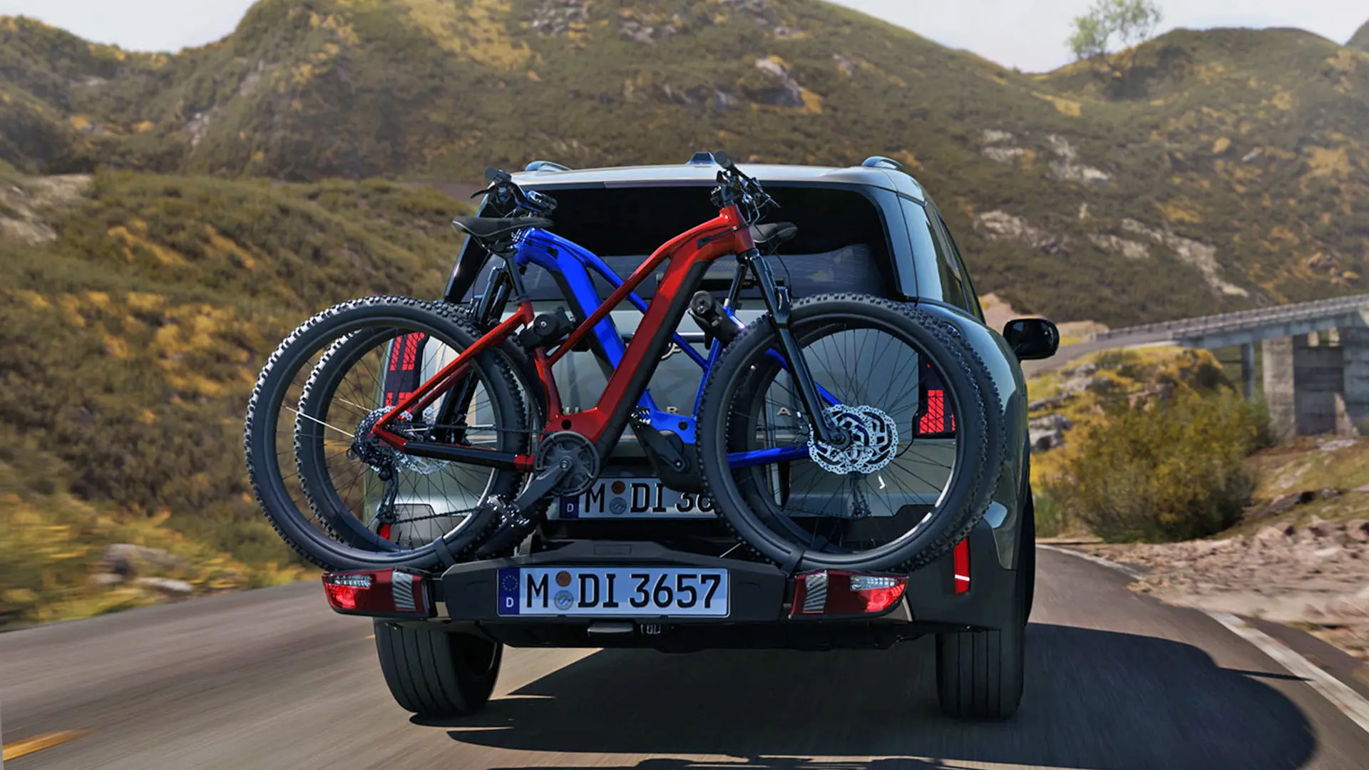 MINI Countryman em Smokey Green equipado com um sistema de suporte traseiro para bicicletas e duas bicicletas de montanha encaixadas, vista traseira, a conduzir ao longo de uma estrada de montanha.