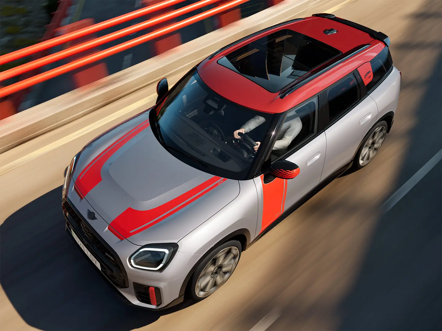 MINI Countryman John Cooper Works em Melting Silver com tampa do retrovisor exterior, decorações laterais e no capot, tejadilho e winglet em Chili Red, circulando pelas ruas da cidade – vista frontal.