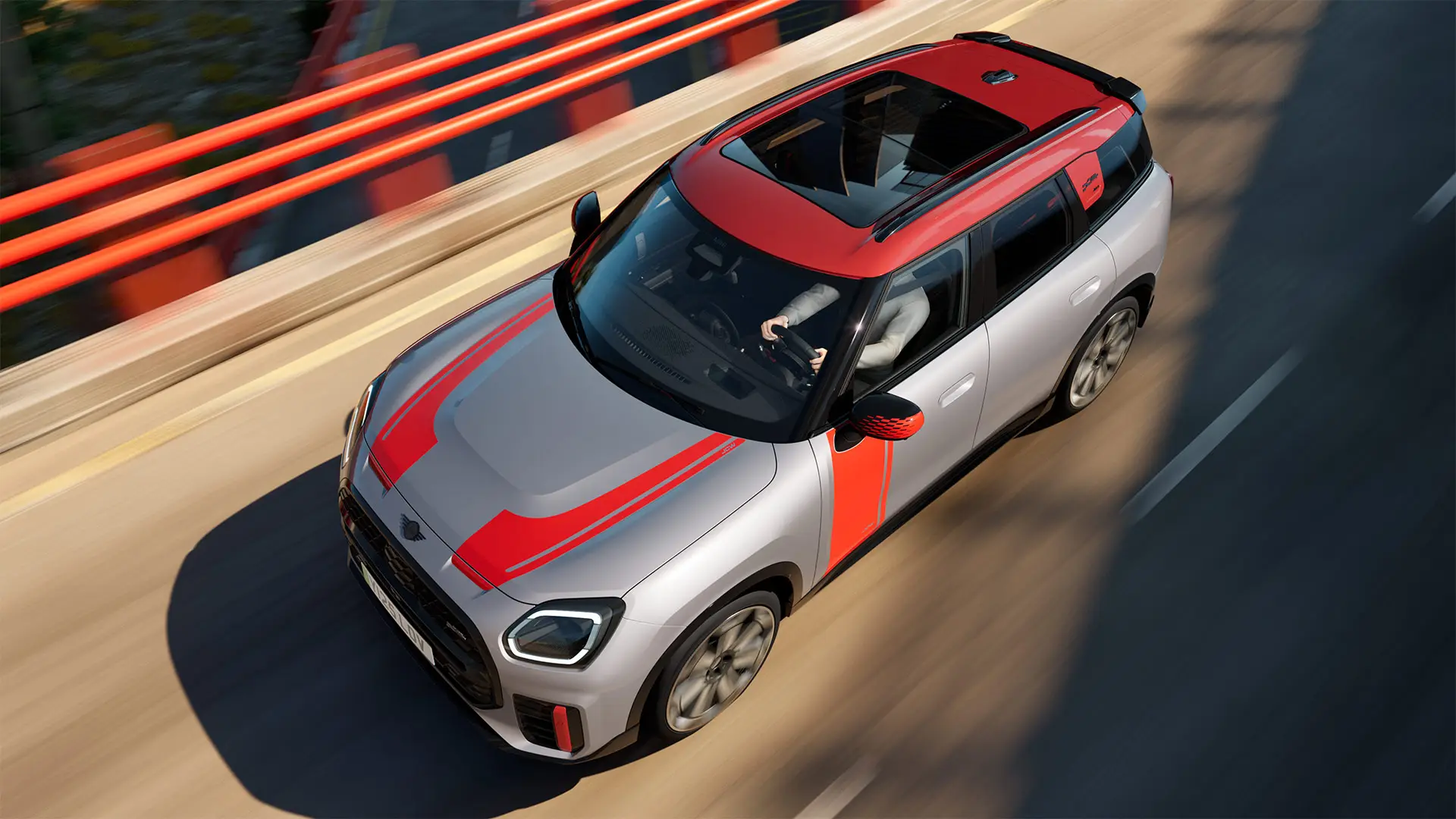 MINI Countryman John Cooper Works em Melting Silver com tampa do retrovisor exterior, decorações laterais e no capot, tejadilho e winglet em Chili Red, circulando pelas ruas da cidade – vista frontal.
