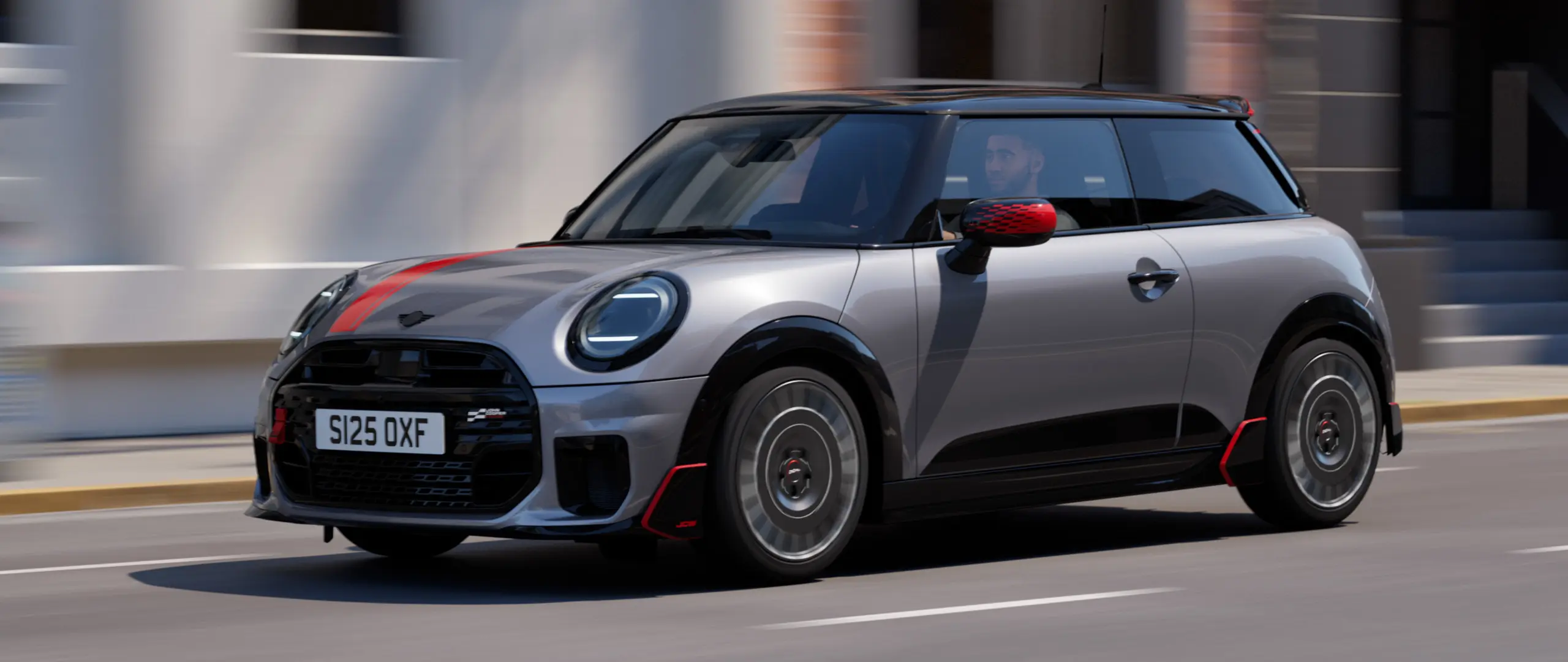 MINI Cooper de 3 portas em Melting Silver com tampa do retrovisor exterior John Cooper Works e decoração de capô em Chili Red, spoiler do tejadilho e winglet em Preto, circulando pelas ruas da cidade – vista frontal.
