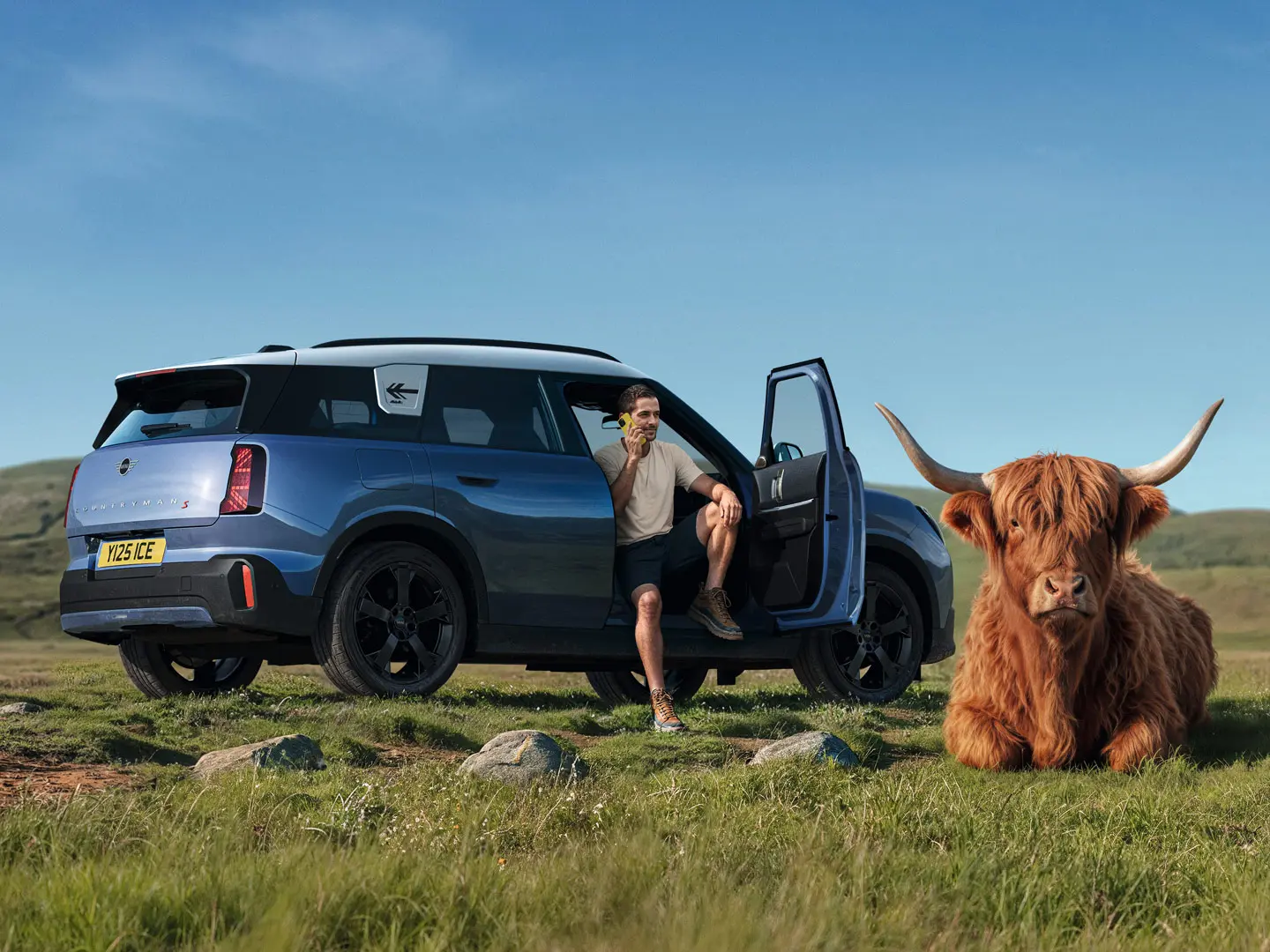 Homem sentado no seu MINI Countryman S com a porta aberta e as pernas no exterior, a falar ao telemóvel com a Assistência em Estrada MINI, enquanto um touro Highland repousa por perto.