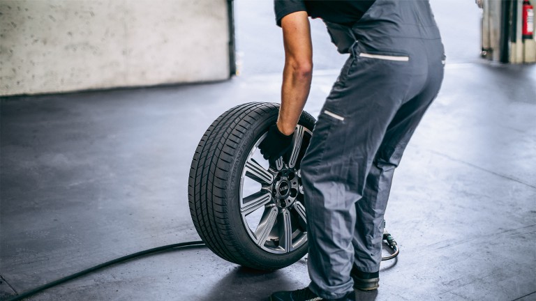 Colaborador do MINI Service num fato-macaco cinza a substituir um pneu - perspetiva traseira.